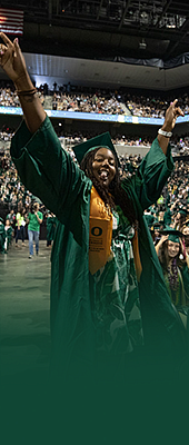 Graduating student walking back to their seat in Matthew Knight Arena, throwing their hands in the air in celebration. The arena is filled with other students in their graduation regalia and the surrounding seats are filled with supporters.