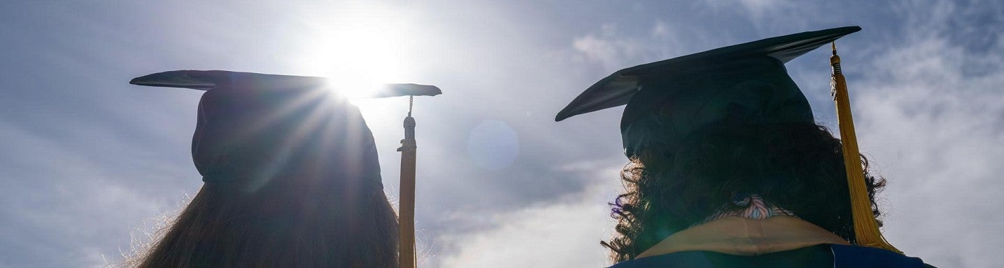 two students facing the sun creating a silhoutte while they wear their cap and gown