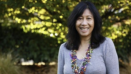 Lanch McCormick smiling wearing a long beaded necklace and a long-sleeve, light grey shirt. There is a slightly blurred tree in the background. 