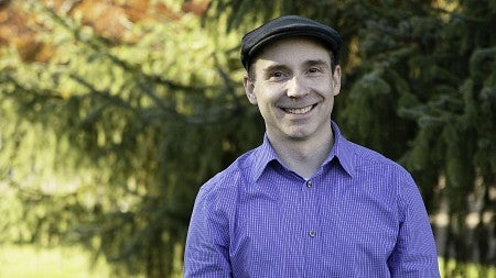 Kevin Hatfield smiling, wearing a black flat cap hat, and a blue and white pinstriped button up shirt. There is a slightly blurred tree in the background. 