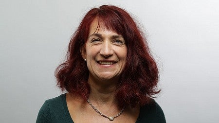 Lara Fernandez smiling, wearing a dark green blouse and a silver chain necklace with a small pearl pendant. She is standing with a white wall in the background.