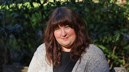 Amy Hughes Giard smiling in the sunlight while wearing a black blouse and light grey cardigan. She is sitting in front of a slightly blurred background of bushes. 