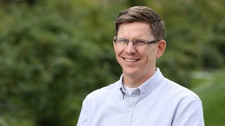 Grant Schoonover smiling, wearing rimless glasses and a light blue button up shirt. He is standing in front of a slightly blurred background of bushes and trees.