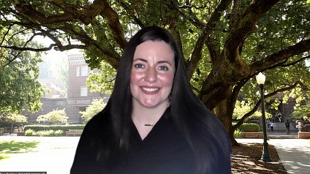 Christabelle Dragoo smiling, wearing a small gold chain necklace and black button up shirt. She is standing in front of a large tree in front of the Knight Library.