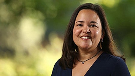 Lori Manson smiling, wearing a black blouse and a matching black necklace with a small pendant. She is standing in front of a blurred background of bushes and trees. 