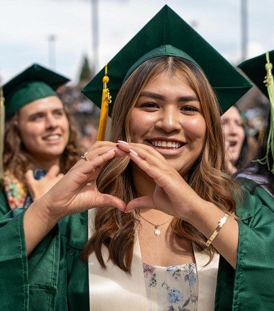 student wearing green graduation regalia using hands to form an 'o'