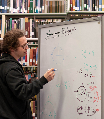 student writing on whiteboard solving a math equation wearing a black hoodie, glasses, and hair in a ponytail