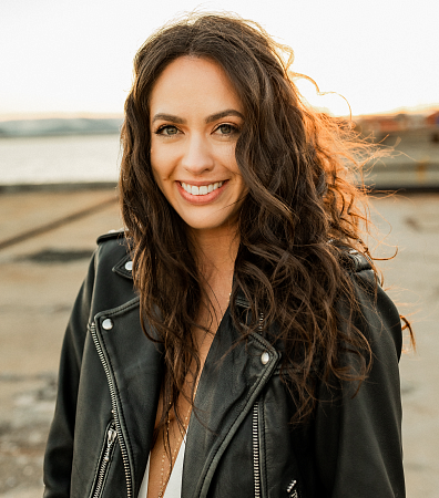 PathwayOregon alumni Madi Steele headshot taken place outside in a field. She is wearing a black leather jacker and her hair brown and curly.