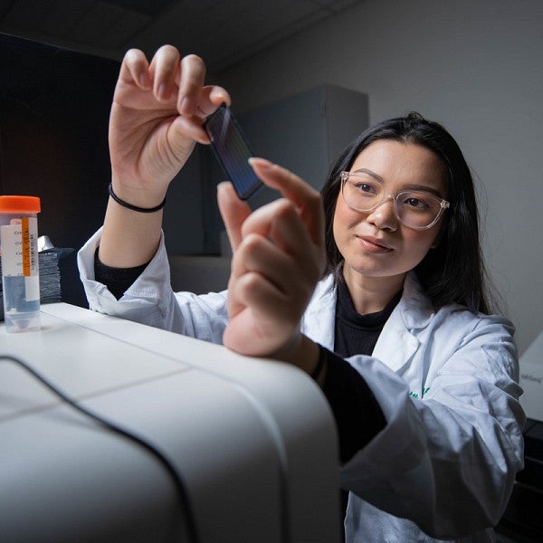 Female student holding up and examining a microscope slide in a research lab. Her hands are resting on a machine. The student is wearing lab glasses and a lab coat.