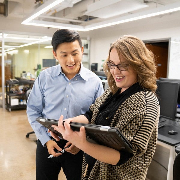 Male staff member is smiling and holding a dry erase marker while slightly leaning towards a female student who is holding a touch screen laptop device and pointing at a problem on the screen. They are standing in front of a dry erase board.