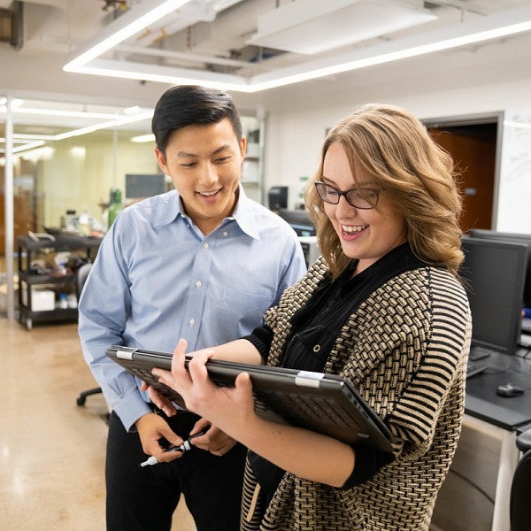 Male staff member is smiling and holding a dry erase marker while slightly leaning towards a female student who is holding a touch screen laptop device and pointing at a problem on the screen. They are standing in front of a dry erase board.   