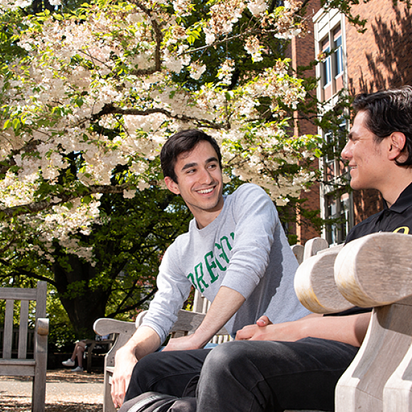 Two male students sitting on a wooden bench outside the Knight Library with a tree in the background that has white flowers blooming.