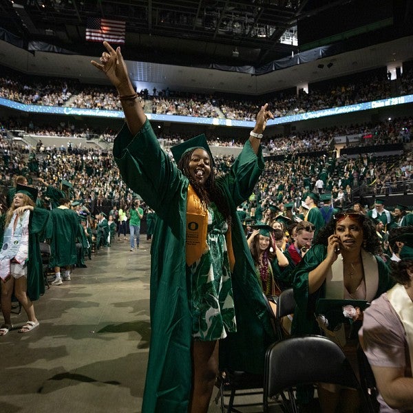 Graduating student walking back to their seat in Matthew Knight Arena, throwing their hands in the air in celebration. The arena is filled with other students in their graduation regalia and the surrounding seats are filled with supporters.