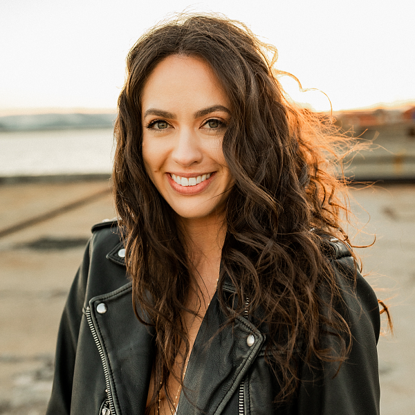 PathwayOregon alumni Madi Steele headshot taken place outside in a field. She is wearing a black leather jacker and her hair brown and curly.