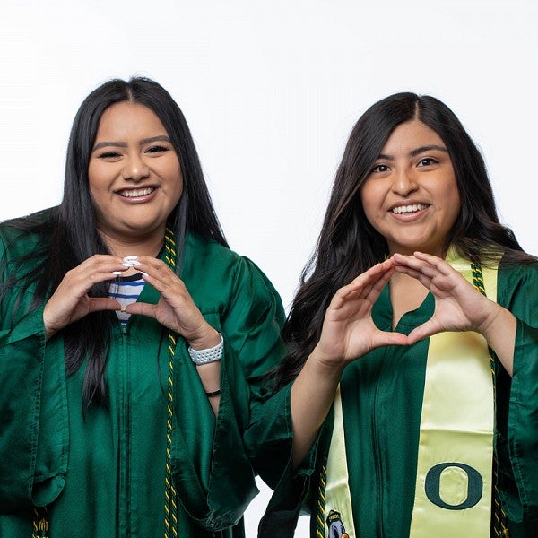 Two female students smiling, wearing UO green graduation gown, and throwing their O