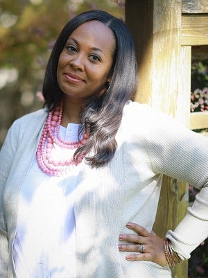 Kimberly Johnson smiling and wearing a white button up shirt with a light beige sweater and pink statement necklace while leaning against a wooden post and her hand placed on her hip.