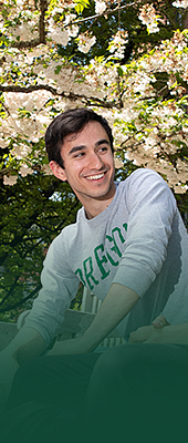 Male student sitting on a wooden bench outside the Knight Library with a tree in the background that has white flowers blooming.