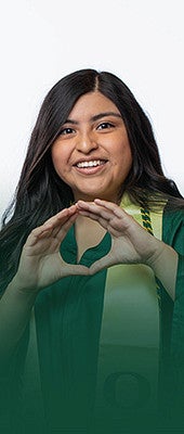 Female student smiling, wearing UO green graduation gown and gold graduation stole and throwing their O at chest level. 