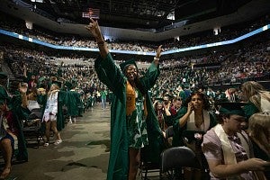 Student standing in Matthew Knight Arena, throwing their hands in the air in celebration of graduation. The arena is filled with other students in their graduation regalia and family members watching from the seats.