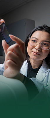 Female student holding up and examining a microscope slide in a research lab. Her hands are resting on a machine. The student is wearing lab glasses and a lab coat.
