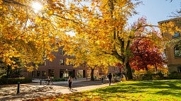 Three buildings on campus slightly blocked by large trees filled with orange and red leaves. There are students walking on the side walks and a student is riding their bike through the road between the buildings. 