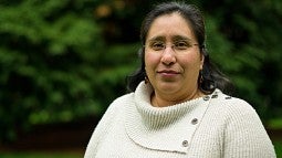 Rosa Chavez smiling, wearing rimless glasses and a light beige sweater with bronze buttons. She is standing in front of a blurred background of bushes and trees.