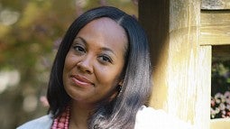 Kimberly Johnson smiling, wearing a white shirt, light beige cardigan and pink necklace while leaning up against a wooden post.