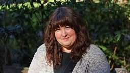 Amy Hughes Giard smiling in the sunlight while wearing a black blouse and light grey cardigan. She is sitting in front of a slightly blurred background of bushes.