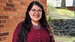 Daisy Martinez smiling wearing black glasses and a maroon button up shirt with a matching statement necklace. She is standing partially in front of a brick wall off to the left and a tree off to the right.