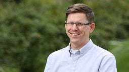Grant Schoonover smiling, wearing rimless glasses and a light blue button up shirt. He is standing in front of a slightly blurred background of bushes and trees.