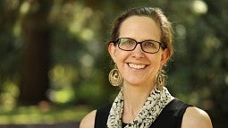 Julie Voelker-Morris smiling, wearing black glasses and gold, circular chandelier earrings. She is wearing a black sleeveless blouse and a patterned scarf that is yellow and black.