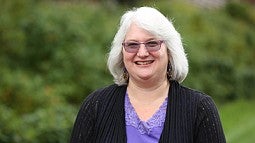Hilary Gerdes Hilary Gerdes smiling wearing purple glasses, a purple shirt with flower embroidery around the neckline and a black sweater. She is standing in front of a blurred background of bushes and trees.