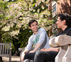 Two male students sitting on a wooden bench outside the Knight Library with a tree in the background that has white flowers blooming.